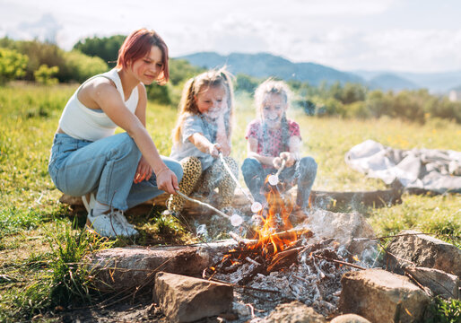 Three sisters cheerfully smiling while they roasting a  marshmallows candies on the sticks over the campfire flame. Happy family or outdoor picnic activities concept image.