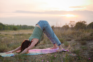 Attractive woman in jeans doing physical exercises on nature background at sunset light. Yoga, stretching, healthy way of life.