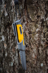 Tourist survival knife with yellow handle, grey sheath, knife hanging on a tree.