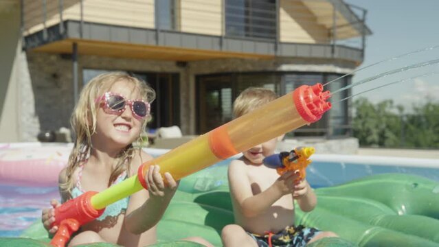 CLOSE UP: Cheerful brother and sister in swimming pool spraying with water guns. Happy children laughing and enjoying water game at backyard pool party. Splashing water for refreshment in summertime.
