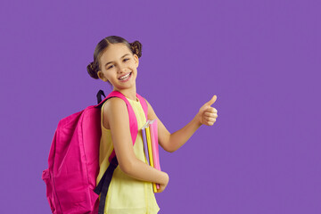 Little girl with backpack and notebooks shows thumb up as sign of approval on purple background....