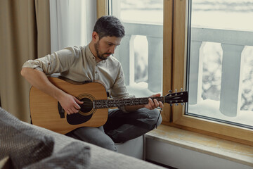 Pensive musician play acoustic guitar composing music, sitting near window at home. Focused...
