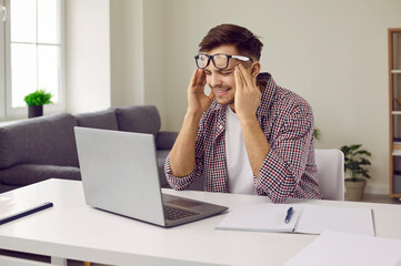 Tired stressed man sitting at working desk with laptop computer, suffering from terrible headache, holding his head, making upset face expression and rubbing his temples. Stress, pain, fatigue concept