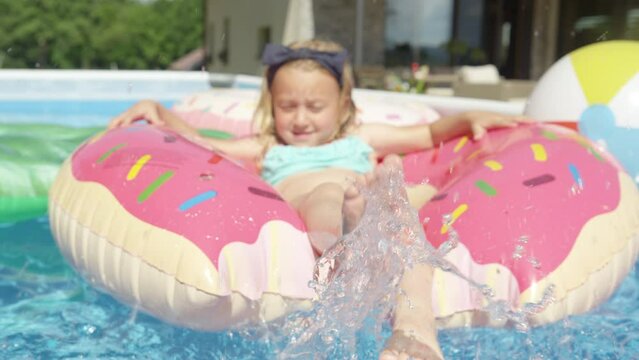 CLOSE UP: Adorable girl floating in pool on inflatable donut and splashing water. Cheerful kid enjoying while having fun in home swimming pool. Outdoor family activities for hot sunny summer days.