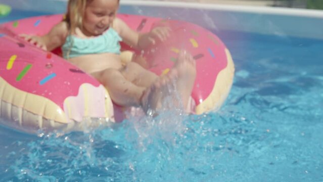 CLOSE UP: Little girl legs splashing water from a floatie donut in swimming pool. Cute girl having lots of fun while making water splashes with her legs. Cheerful kid enjoying summer in backyard pool.