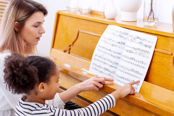 Fototapeta na wymiar Little girl reading song and practicing playing the piano, pupil learning how to play musical instrument on lesson. Lovely woman teacher explain small black child how to play notes. Hobby and activity