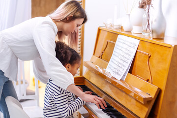 Little black girl pupil is playing, learning and practicing the piano. Music abilities for kids. Hobby and activity lessons for the children. Hand of an experienced pianist woman helping young student