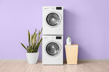 Modern drying, washing machines, basket and houseplant near violet wall in room