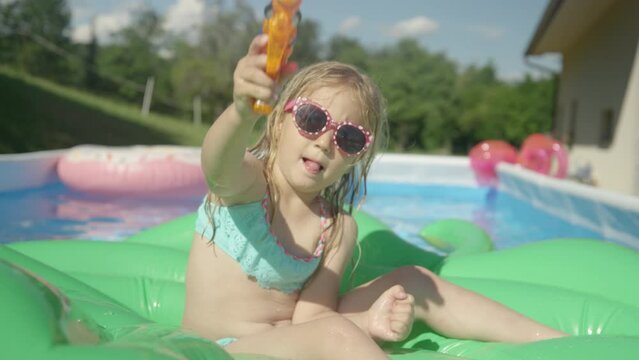CLOSE UP: Little girl sitting on a floatie with water gun splashing into camera. Playful little girl floating in backyard swimming pool and having fun with water blaster spraying directly into camera