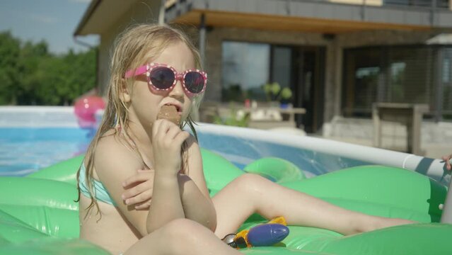 CLOSE UP: Adorable girl floating in pool and eating ice cream on a hot summer day. Cute little girl enjoying chocolate ice cream bar in backyard garden pool. Complete refreshment on a hot summer day.