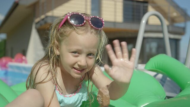 CLOSE UP: Sweet little girl with ice cream bar in the pool waving towards camera. Adorable girl enjoying chocolate ice cream bar in backyard garden pool. Complete refreshment on a hot sunny summer day