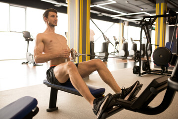 Shirtless muscular athlete sitting and pulling the weight on himself while training on rowing...