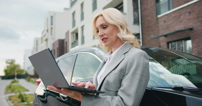 Beautiful caucasian middle-age woman in formal clothes standing in front of the black car, using laptop on the side of the road in city