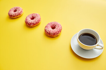 Breakfast concept: close-up of full coffee cup on plate and doughnuts with pink topping placed in a row, yellow background