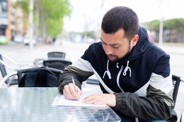 Focused bearded guy sitting at table in outdoor cafe on spring day, filling out papers..
