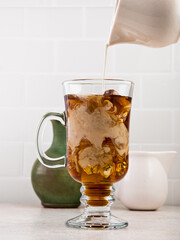 Iced coffee with cream pouring into a clear tall glass with a white tile wall in background