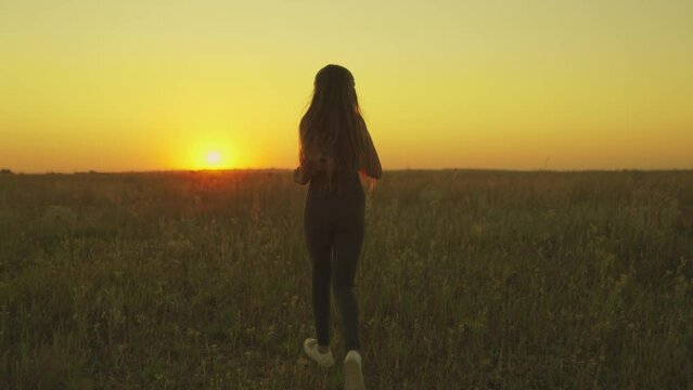 young girl runs across field sunset headphones. silhouette girl rays sunlight listens music. healthy image women health. morning run outdoors cardio. exercises weight loss. long-haired sun girl.
