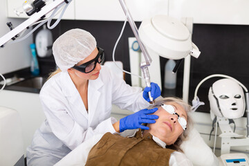 Focused skilled female cosmetologist performing hardware laser treatment to resurfacing and tightening facial skin to senior woman at cosmetology clinic