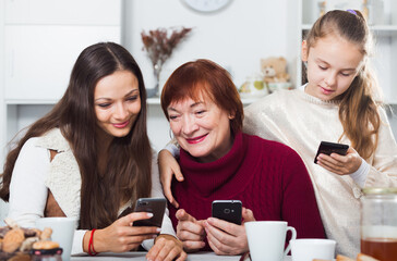 Happy grandmother, mother and daughter using mobile gadgets at table at home