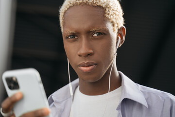 Closeup portrait of confident African American man with stylish curly hair wearing casual clothing holding mobile phone looking at camera. Hipster guy listening music outdoors 