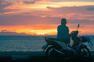 Fototapeta na wymiar Woman sitting on the scooter by the sea and enjoying the colorful sunset