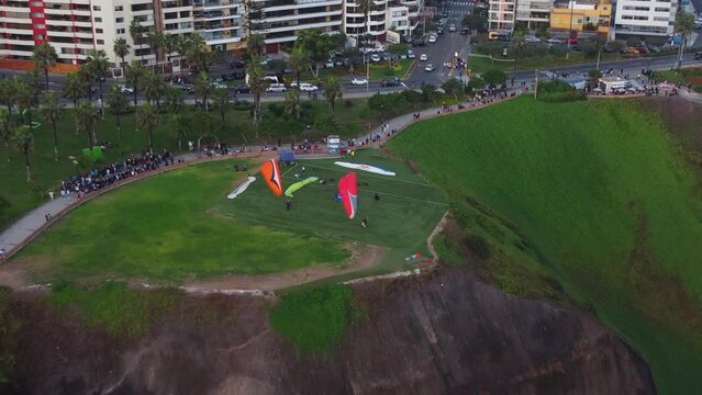 Drone video of paragliders in Lima, Peru. In Miraflores district. Zooming out as paragliders prepare to take off from the green hill. Many people, trees, grass andbuildings can be seen in background
