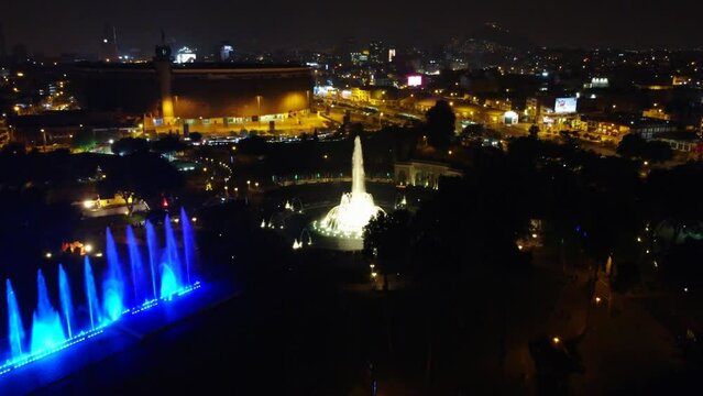 Drone video of colorful water fountain in Lima, Peru. Slowly flying towards main circular fountain. Many city lights can be seen in background.