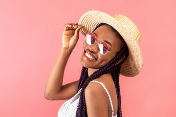 Cheerful black lady with braids wearing stylish dress and sunglasses, touching straw hat and smiling at camera - obrazy, fototapety, plakaty