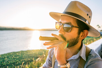 Latin young man wearing glasses, hat and suspenders smoking a cigar in a bar on the shore of a lake...