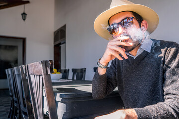 Young Nicaraguan man wearing a sweater hat and sunglasses smoking a cigar in the living room of a...