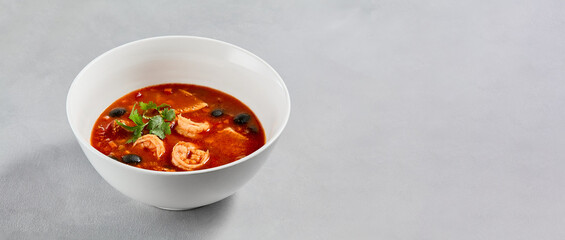Fish tomato soup with shrimps and black olives. Seafood soup with prawns in white ceramic bowl on concrete background. Fish dish with seafood. Tomato soup in minimal composition.