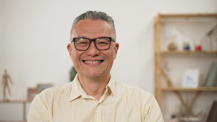 shoulder shot portrait of a cheerful Chinese elderly male in casual wear smiling at the camera in a modern bright home interior