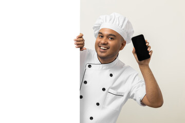 Happy Young handsome asian man chef in uniform looks out from behind an empty whiteboard holding...