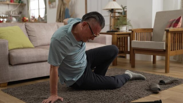 Asian elderly man trying to slowly get up after he slipped and fell on floor in the living room at home. he feels great pain in lower back while using stick to support himself