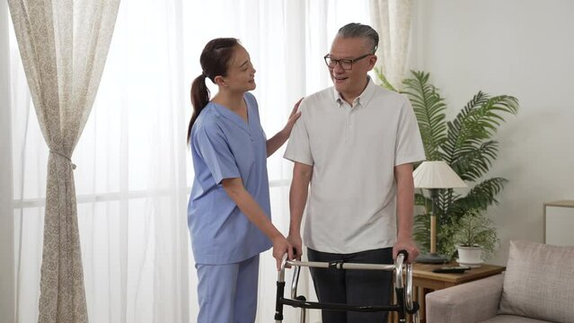 cheerful asian older man under recovery process is talking happily with female nursing aide by window in a bright home interior. he stands with the support of walker