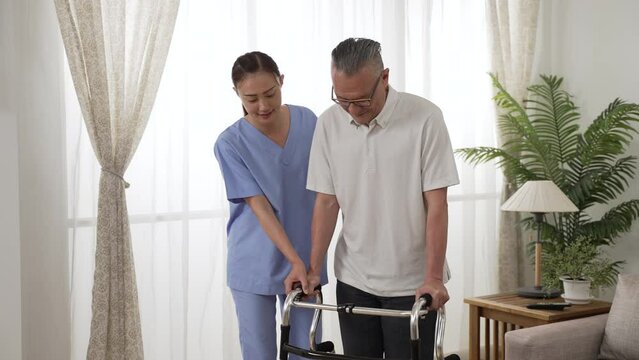 asian senior man undergoing rehabilitation with the help of woman resident care attendant. he uses a walker to practicing walking slowly by the window at home