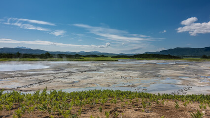 Fototapeta na wymiar There are hot springs in the valley of the caldera of an extinct volcano. Steam rises above the water. Traces of bear paws are visible on the sandy soil. Mountains and blue sky. Kamchatka. Uzon