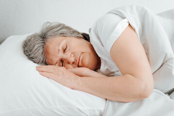 Senior caucasian woman sleeps in bed in bedroom lying on her side and arms folded. Elderly woman morning dream, close-up