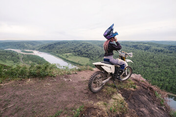 Woman biker wearing helmet and pink dreadlocks sitting on her dirt motorcycle on mountain top above Beautiful view