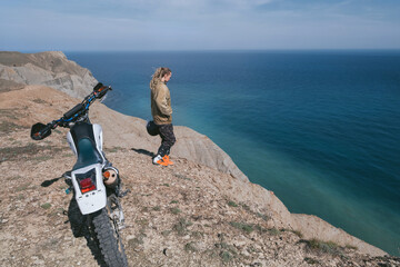 Female rider relaxing near dirt motorcycle on stunning blue sea cliff top