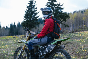 Female wearing helmet sitting on enduro dirt motorcycle on field with yellow flowers and pine forest