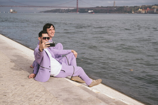 Happy gay couple seating on city beach, enjoy romantic summer day, doing selfie by mobile phone