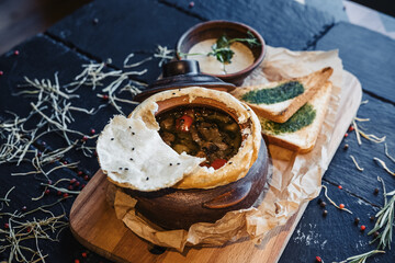 Roast with meat, potatoes in clay pot under dough, crispbreads and cream sauce on wooden board