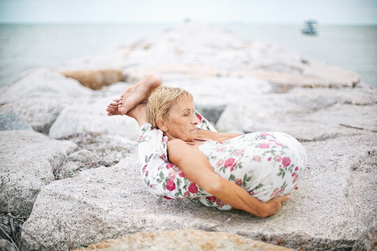 Elderly woman yoga master holding her legs behind her head in yoga nidrasana pose on white stones of sea shore