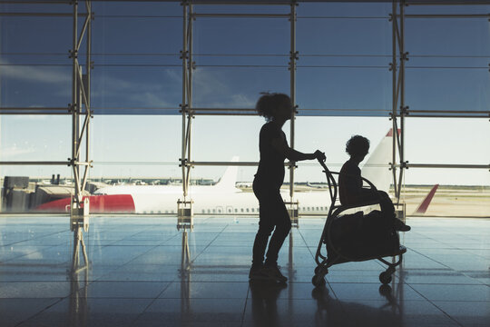 Silhouette of woman carrying child in luggage cart in airport terminal in front of large windows overlooking the planes