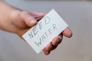 Need water. Words written in jagged letters. A man's hand holds a white paper rectangle with text against a gray background.