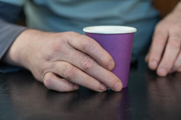 A grown man holds a lilac glass in his hands. A paper cup for hot drinks. Eco-friendly utensils concept. Man sitting at a black table indoors. Close-up. Selective focus.