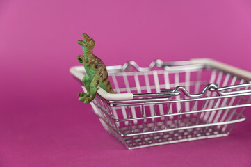 Dinosaur and shopping cart against a purple background. The dinosaur sits on a white handle. Green miniature of an animal of prey. Selective focus.