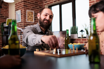 Smiling man moving chess piece on board while sitting at table with close friends. Multiethnic people in living room at home playing board games while consuming snacks and beverages.