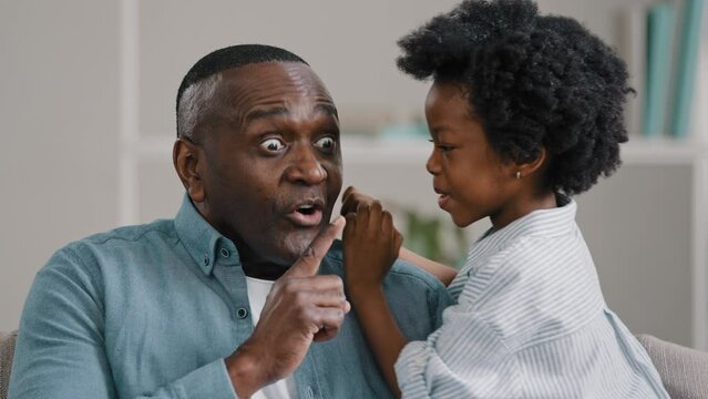 Little african american daughter whispering secret adult dad in ear telling gossip surprised dad carefully listens confidential information making gesture silence secret puts index finger to mouth